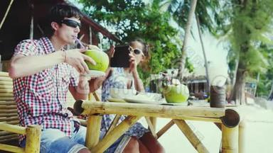 一对戴着太阳镜的年轻夫妇坐在海边的一家漂亮的咖啡馆里。 女孩用手机给男友拍照